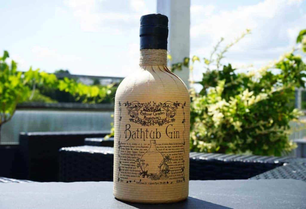 Auf einem Tisch im Freien steht eine Flasche „Bathtub Gin“ mit einem Etikett im Vintage-Stil, umgeben von Grünpflanzen und schwarzen Korbmöbeln, im Hintergrund sind Wasser und ein blauer Himmel zu sehen.