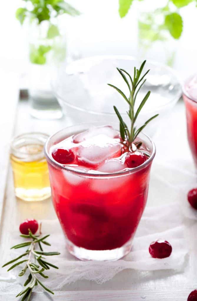 Auf einem Tablett steht ein Glas mit rotem Cranberry-Getränk, Eis, ganzen Cranberries und einem Rosmarinzweig als Garnitur. Daneben stehen ein kleines Glas Honig, weiterer Rosmarin und im Hintergrund verschwommenes Grün.