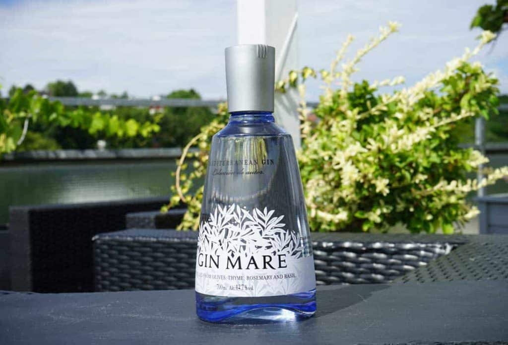 Auf einem grauen Tisch im Freien steht eine blaue Glasflasche Gin Mare, im verschwommenen Hintergrund sind grüne Pflanzen und Wasser zu sehen.