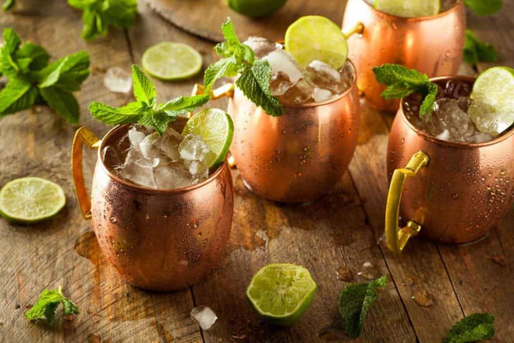 Drei Kupferbecher, gefüllt mit zerstoßenem Eis, Limettenschnitzen und Minzezweigen, stehen auf einem rustikalen Holztisch, auf dem Limettenscheiben und Minzblätter verstreut liegen.
