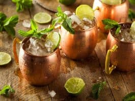 Gin Mule – Gin Cocktail Drei Kupferbecher, gefüllt mit zerstoßenem Eis, Limettenschnitzen und Minzezweigen, stehen auf einem rustikalen Holztisch, auf dem Limettenscheiben und Minzblätter verstreut liegen.