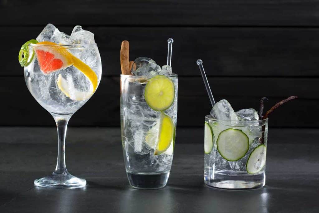 Drei klare Cocktails mit Eis, garniert mit Zitrusscheiben, Gurke, Zimtstange und Vanilleschote, in verschiedenen Gläsern vor einem dunklen Hintergrund.