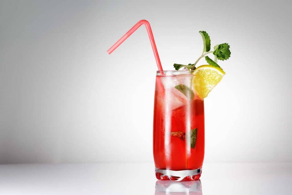 Ein großes Glas mit einem roten Eisgetränk, garniert mit Minzblättern, einer Limettenscheibe und einem roten Strohhalm, vor einem hellen Hintergrund.