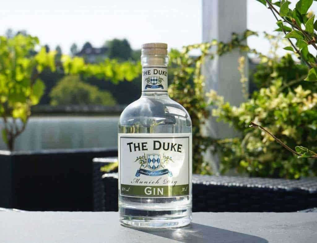 Auf einem Tisch im Freien steht eine Flasche „The Duke Munich Dry Gin“, im Hintergrund sind an einem sonnigen Tag Grünflächen, ein See und verschwommene Bäume zu sehen.