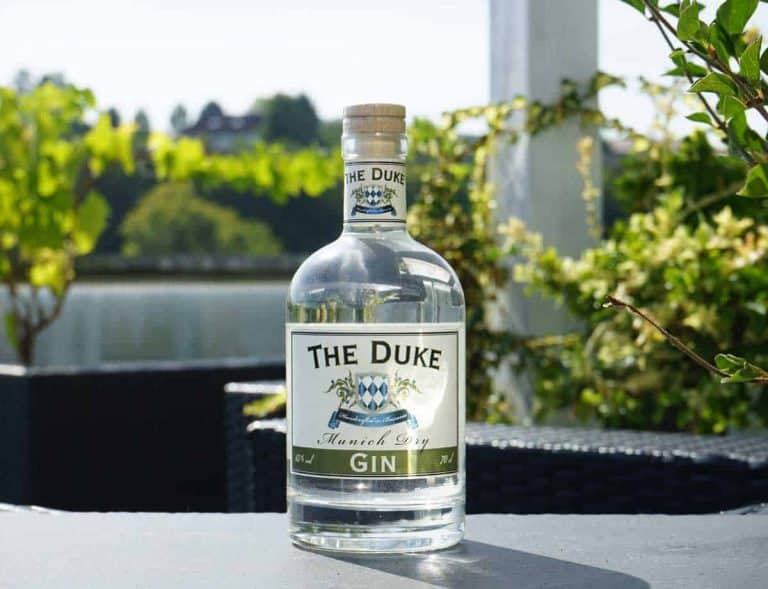 Auf einem Tisch im Freien steht eine Flasche „The Duke Munich Dry Gin“, im Hintergrund sind an einem sonnigen Tag Grünflächen, ein See und verschwommene Bäume zu sehen.