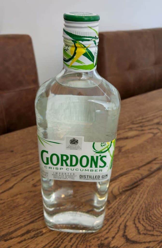 Auf einem Holztisch steht eine Flasche „Gordons Crisp Cucumber Distilled Gin“, im Hintergrund sind braune Stühle zu sehen.