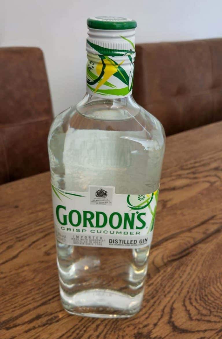 Auf einem Holztisch steht eine Flasche „Gordons Crisp Cucumber Distilled Gin“, im Hintergrund sind braune Stühle zu sehen.