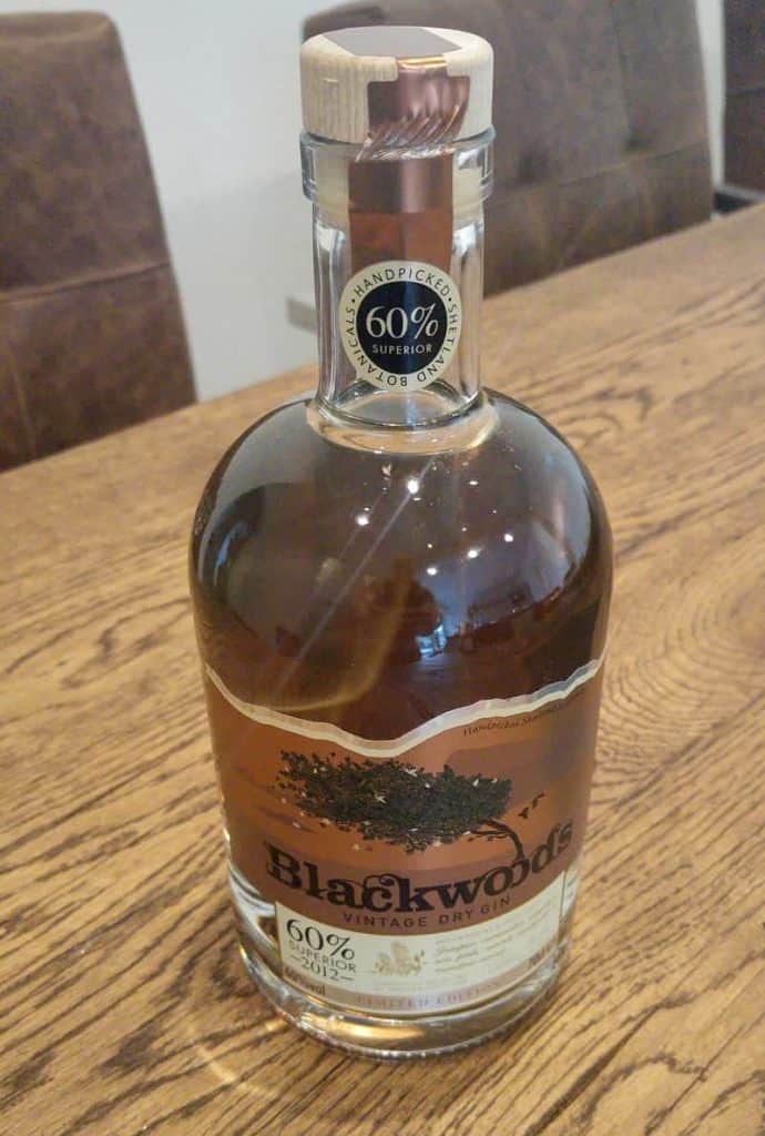 Auf einem Holztisch steht eine Flasche „Blackwoods Vintage Dry Gin“ mit einem Holzverschluss. Auf dem Etikett ist ein Alkoholgehalt von 60 % angegeben und eine Baumillustration abgebildet.