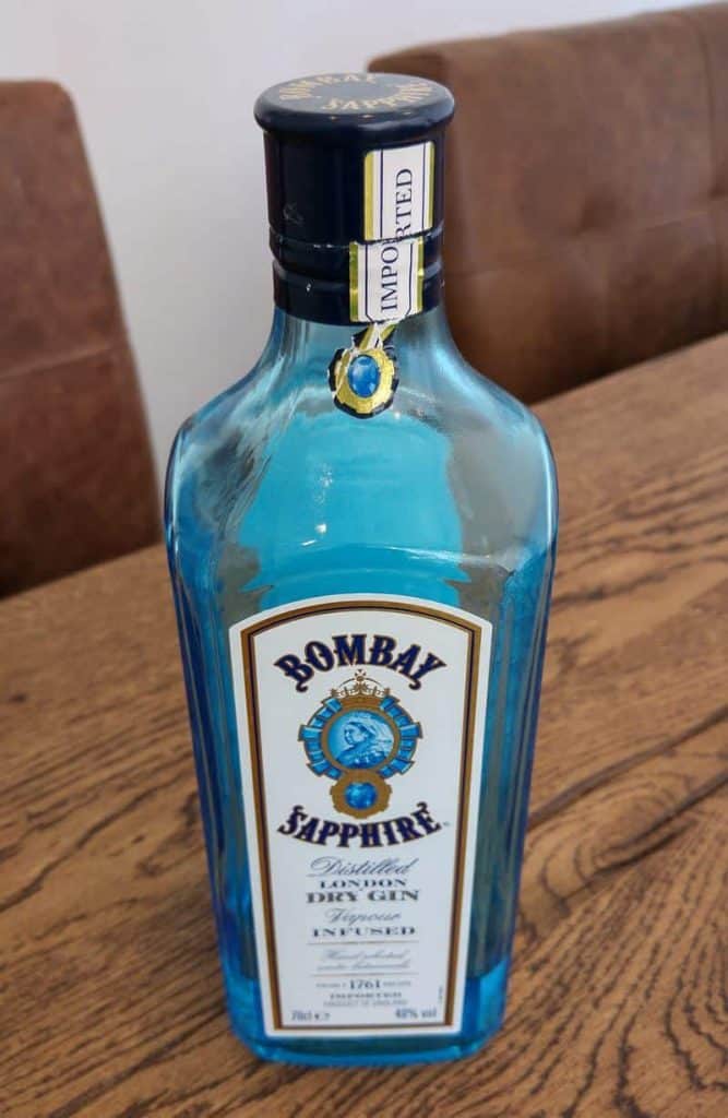 Auf einem Holztisch steht eine blaue Glasflasche „Bombay Sapphire Distilled London Dry Gin“, im Hintergrund ist ein brauner gepolsterter Stuhl zu sehen.