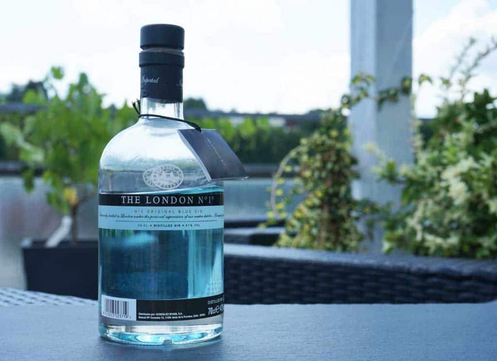 Auf einem Tisch im Freien steht eine Flasche „The London No. 1 Original Blue Gin“, im Hintergrund sind Grünpflanzen und Terrassenmöbel zu sehen.
