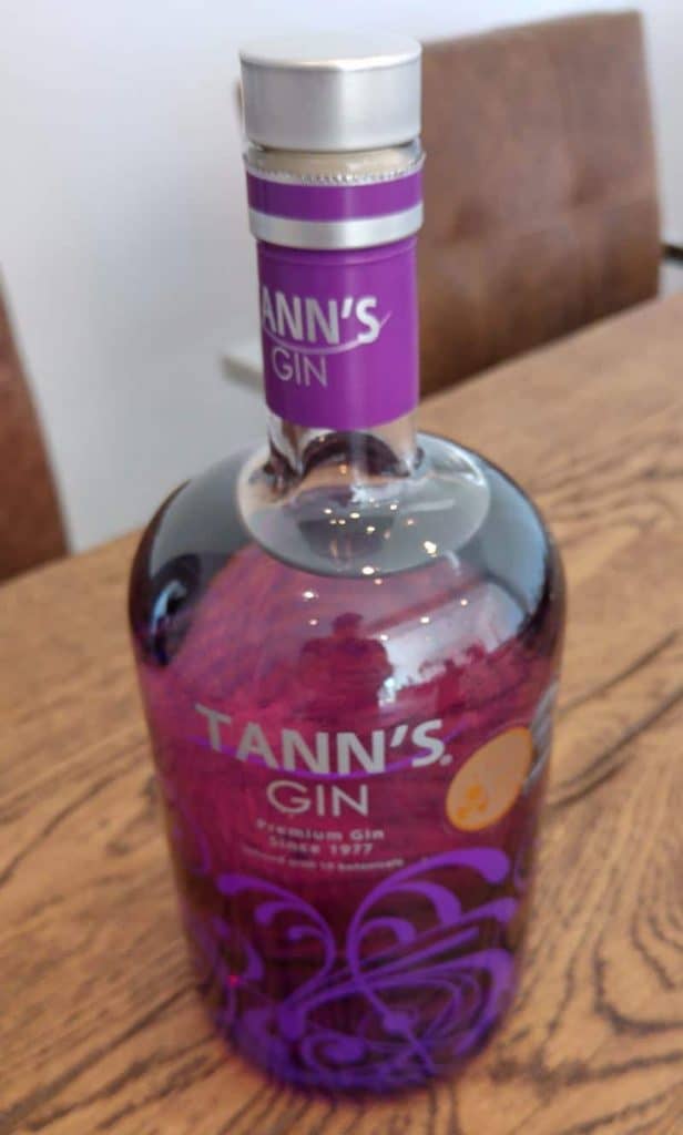 Auf einem Holztisch steht eine Flasche Tanns Gin mit einem violetten Etikett. Im Hintergrund sind zwei braune Stühle zu sehen.