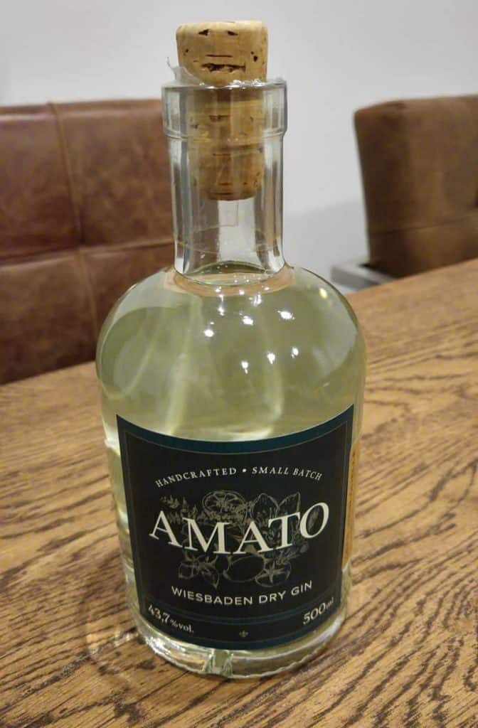 Eine klare Glasflasche „Amato Wiesbaden Dry Gin“ mit Korkverschluss, die auf einem Holztisch steht. Das Etikett ist dunkel und mit weißer und goldener Schrift sowie dekorativen Elementen versehen.