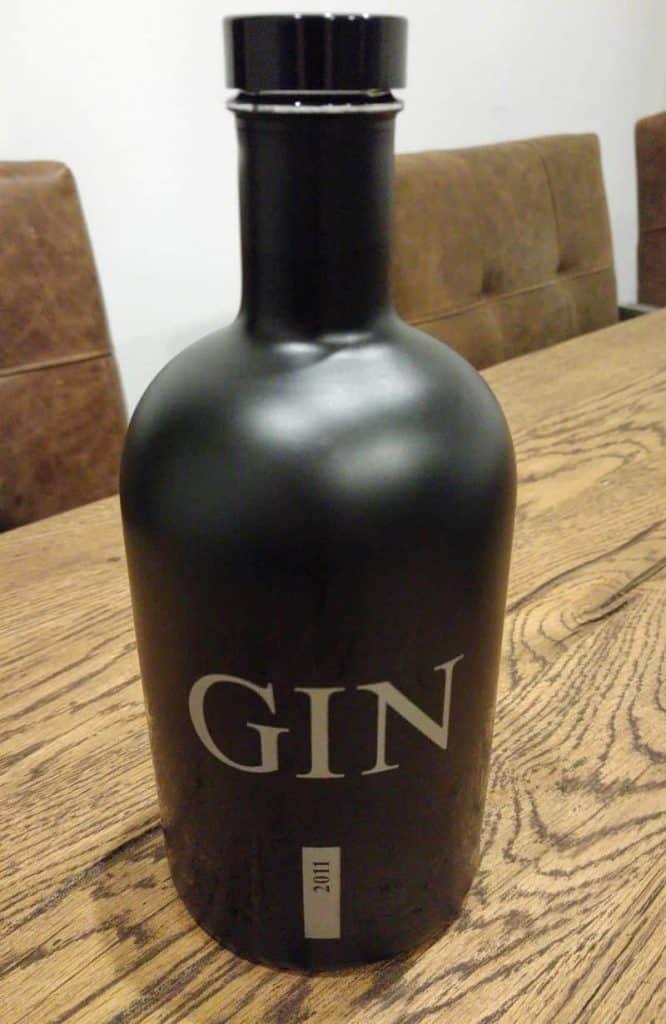 Auf einem Holztisch steht eine mattschwarze Flasche mit der Aufschrift „GIN“ in fetten weißen Buchstaben, im Hintergrund sind braune gepolsterte Stühle zu sehen.
