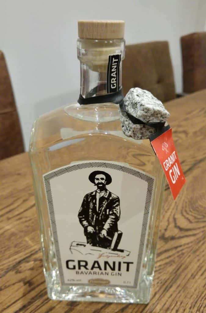 Eine Flasche „Granit Bavarian Gin“ mit einem Etikett, auf dem ein Mann in altmodischer Kleidung abgebildet ist. Am Flaschenhals ist mit einer schwarzen Schnur und einem roten Anhänger ein Granitstein befestigt. Die Flasche steht auf einem Holztisch.