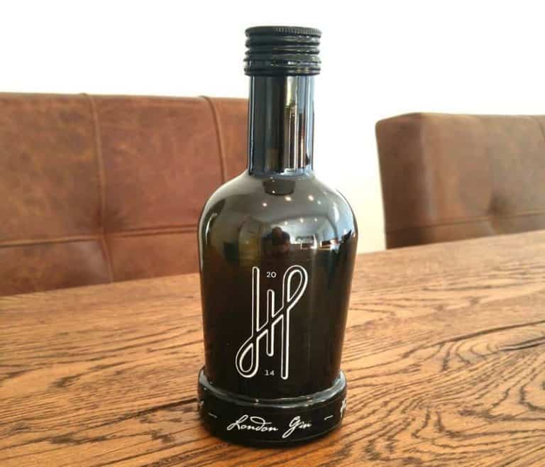 Auf einem Holztisch steht eine schwarze Glasflasche mit der Aufschrift „London Gin“, im Hintergrund sind braune gepolsterte Stühle zu sehen. Die Flasche hat ein modernes weißes Logo und einen Schraubverschluss.