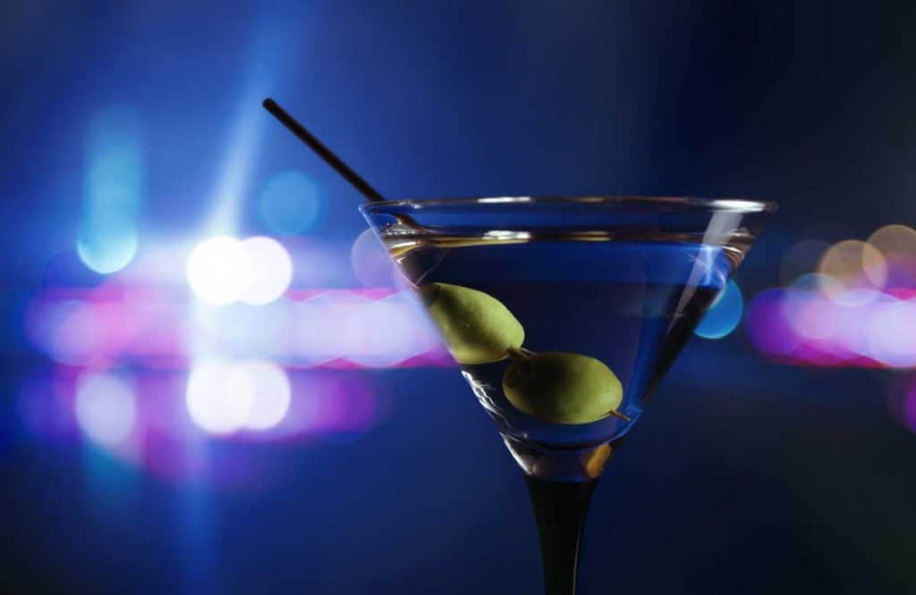 Ein Martini-Glas mit zwei grünen Oliven und einem schwarzen Rührstäbchen vor einem verschwommenen Hintergrund aus blauen und violetten Lichtern.