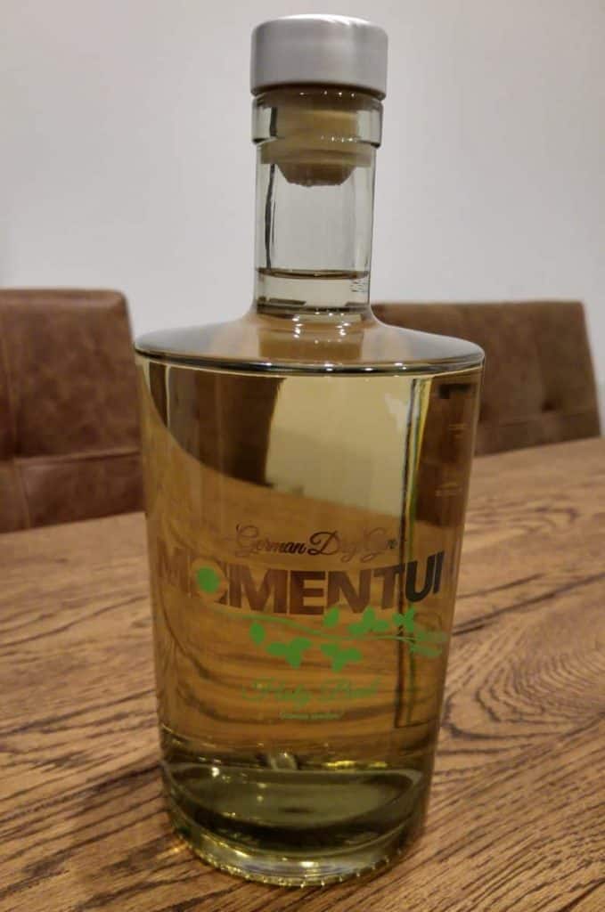Auf einem Holztisch mit braunen Polsterstühlen und einer schlichten Wand im Hintergrund steht eine durchsichtige Flasche mit deutschem Dry Gin, die mit „MOMENTUM“ beschriftet ist.
