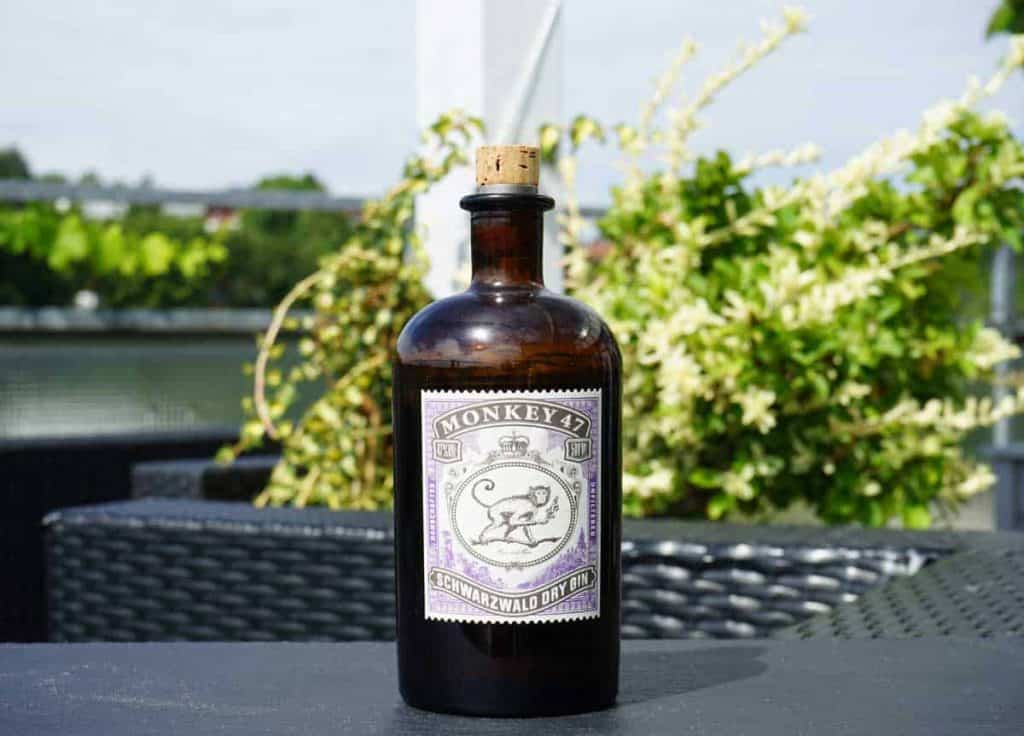 Auf einem dunklen Tisch im Freien steht eine braune Glasflasche „Monkey 47 Schwarzwald Dry Gin“ mit Korkverschluss, im Hintergrund sind grünes Laub und weiße Blüten zu sehen.
