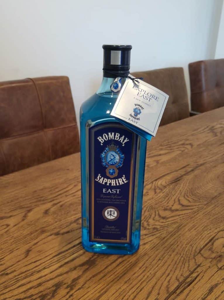 Eine blaue Flasche Bombay Sapphire East Gin mit einem Anhänger am Flaschenhals steht auf einem hölzernen Esstisch, im Hintergrund sind braune Stühle zu sehen.