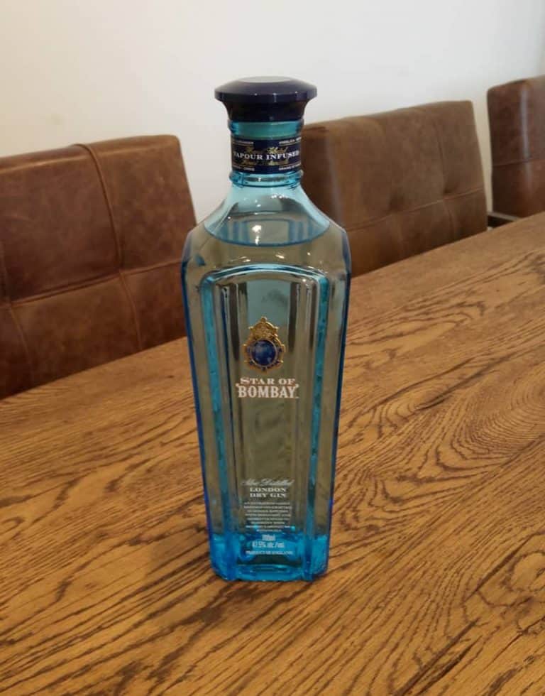 Auf einem Holztisch steht eine blaue Glasflasche mit „Star of Bombay“-Gin, im Hintergrund sind braune Polstersessel und eine weiße Wand zu sehen.