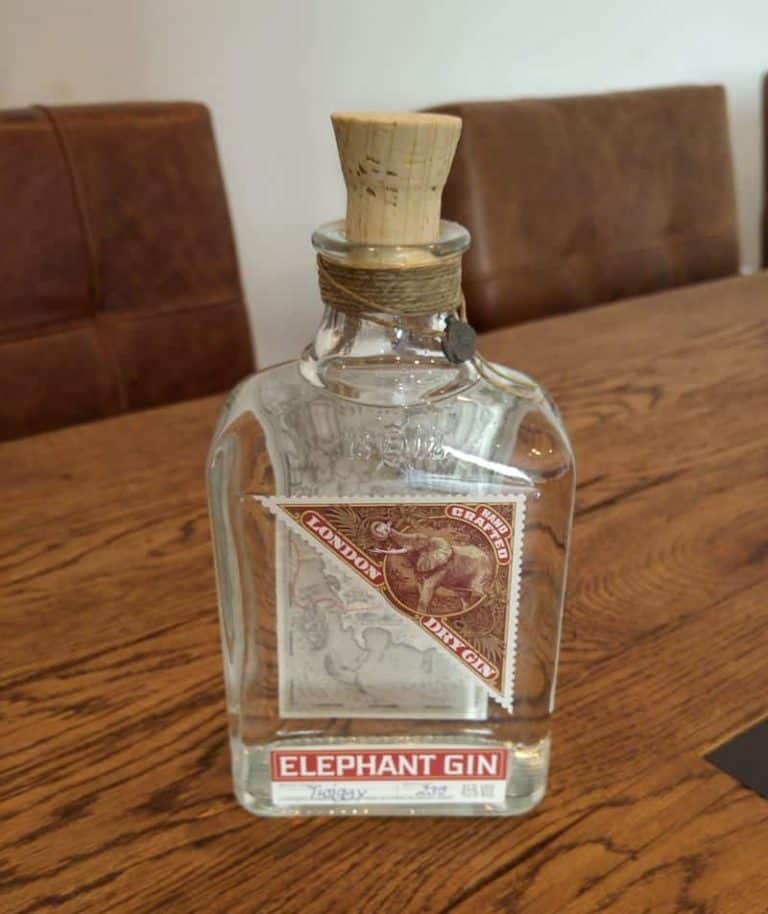 Auf einem Holztisch steht eine Flasche „Elephant Gin“ mit Korkverschluss. Auf dem Etikett sind ein Elefant und ein Stempelmotiv abgebildet. Im Hintergrund sind braune Stühle zu sehen.