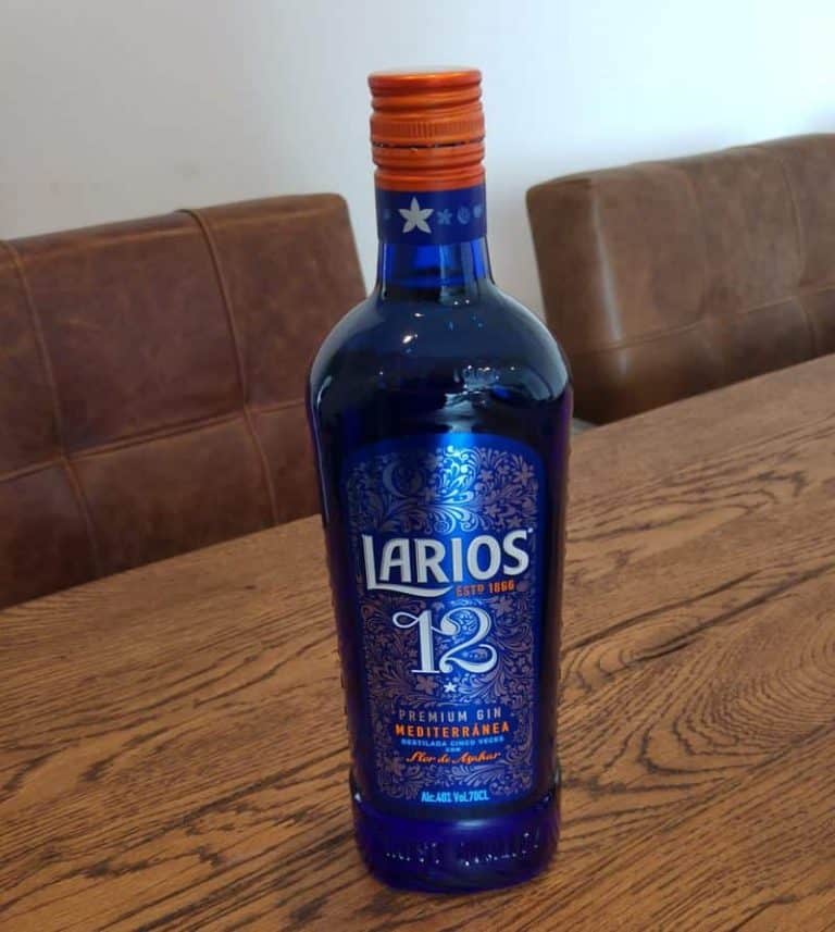 Auf einem Holztisch steht eine blaue Flasche „Larios 12“ Gin, im Hintergrund sind braune Stühle mit Polstern zu sehen.