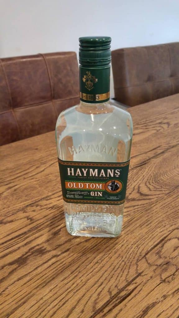 Auf einem Holztisch steht eine Flasche Haymans Old Tom Gin, im Hintergrund ist eine braun gepolsterte Bank zu sehen.