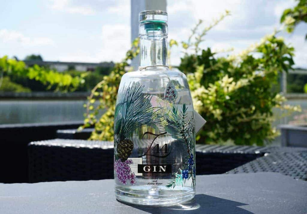 Eine durchsichtige Gin-Flasche mit Botanik-Illustrationen steht auf einem Tisch im Freien, im Hintergrund sind Grünpflanzen und Wasser verschwommen zu sehen.