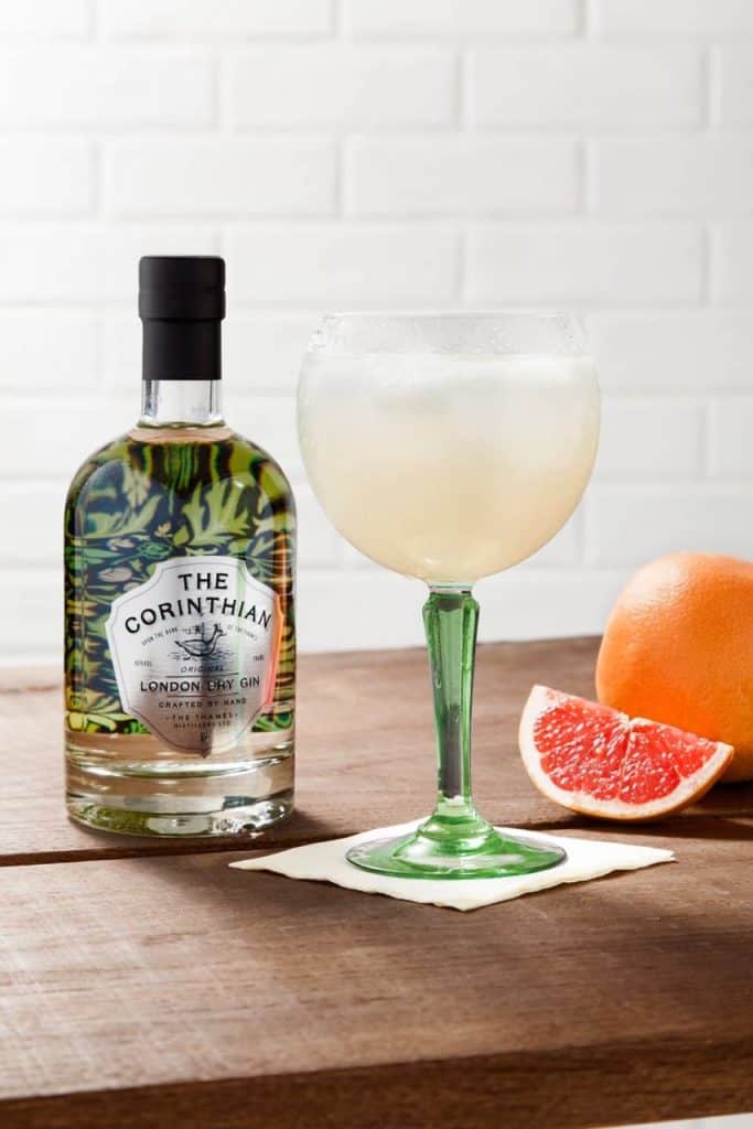 Auf einem Holztisch vor einem Hintergrund aus weißen Ziegelsteinen steht eine Flasche „The Corinthian London Dry Gin“ neben einem Cocktail in einem Glas mit grünem Stiel, einer Grapefruitscheibe und einer ganzen Grapefruit.