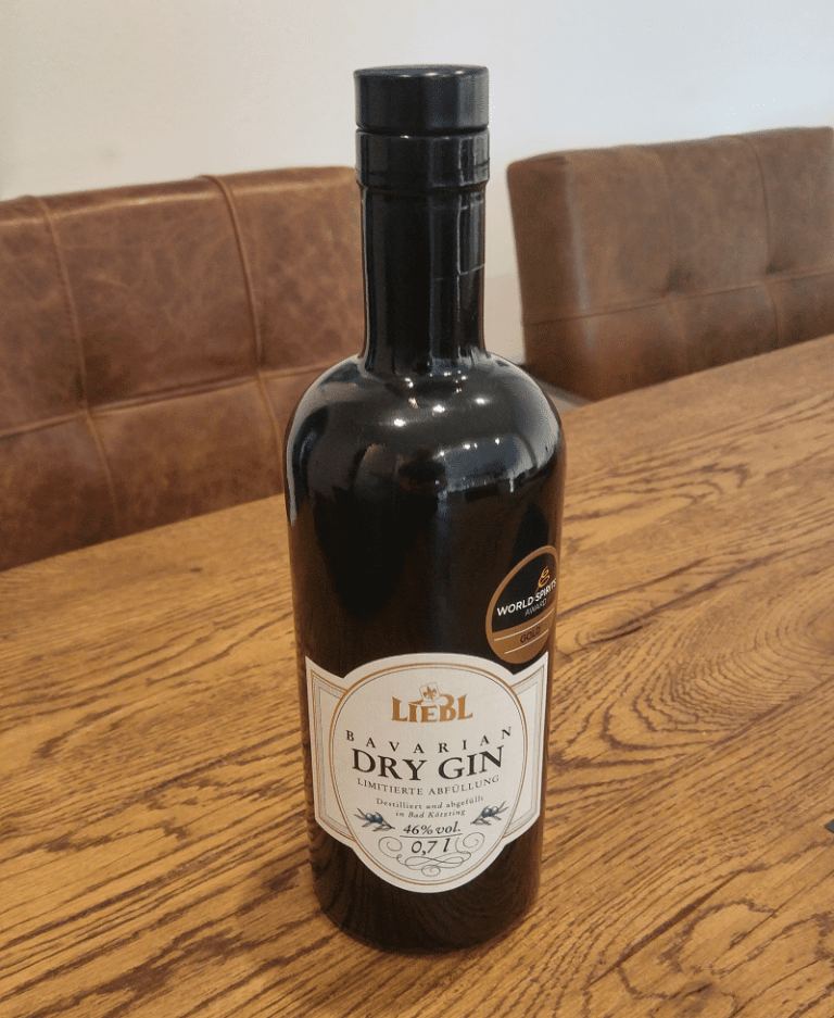 Auf einem Holztisch steht eine Flasche „Liebl Bavarian Dry Gin“, im Hintergrund ist ein brauner gepolsterter Stuhl zu sehen. Die Flasche hat ein weißes Etikett und einen schwarzen Verschluss.