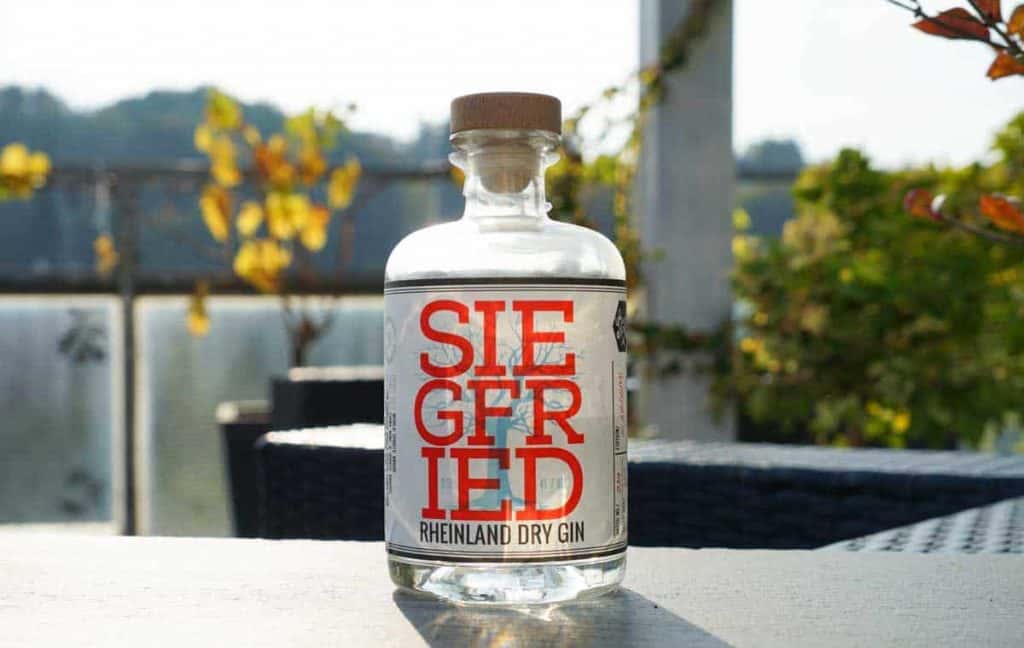 Auf einem Tisch im Freien steht eine durchsichtige Flasche „Siegfried Rheinland Dry Gin“ mit einem Holzverschluss, umgeben von Grün und mit Sonnenlicht im Hintergrund.