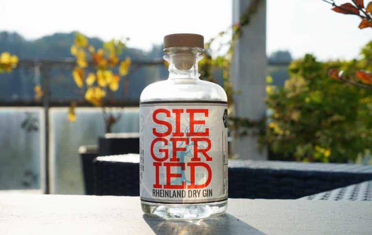 Auf einem Tisch im Freien steht eine durchsichtige Flasche „Siegfried Rheinland Dry Gin“ mit einem Holzverschluss, umgeben von Grün und mit Sonnenlicht im Hintergrund.