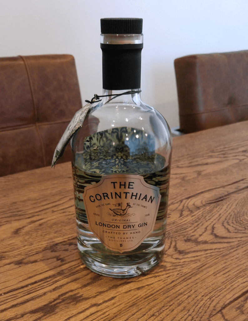 Auf einem Holztisch steht eine Flasche „The Corinthian London Dry Gin“, im Hintergrund sind braune Stühle zu sehen.