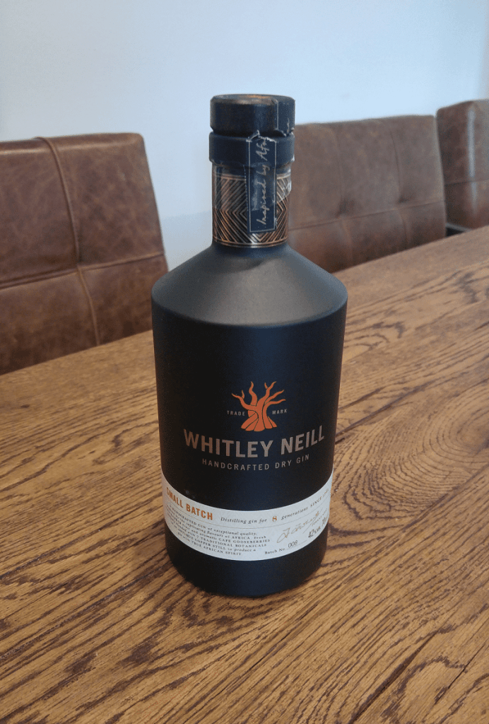 Auf einem Holztisch steht eine Flasche „Whitley Neill Handcrafted Dry Gin“, im Hintergrund sind braune Stühle mit Polstern zu sehen.
