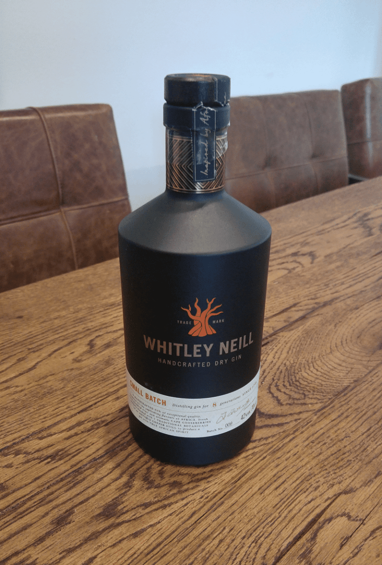 Auf einem Holztisch steht eine Flasche „Whitley Neill Handcrafted Dry Gin“, im Hintergrund sind braune Stühle mit Polstern zu sehen.
