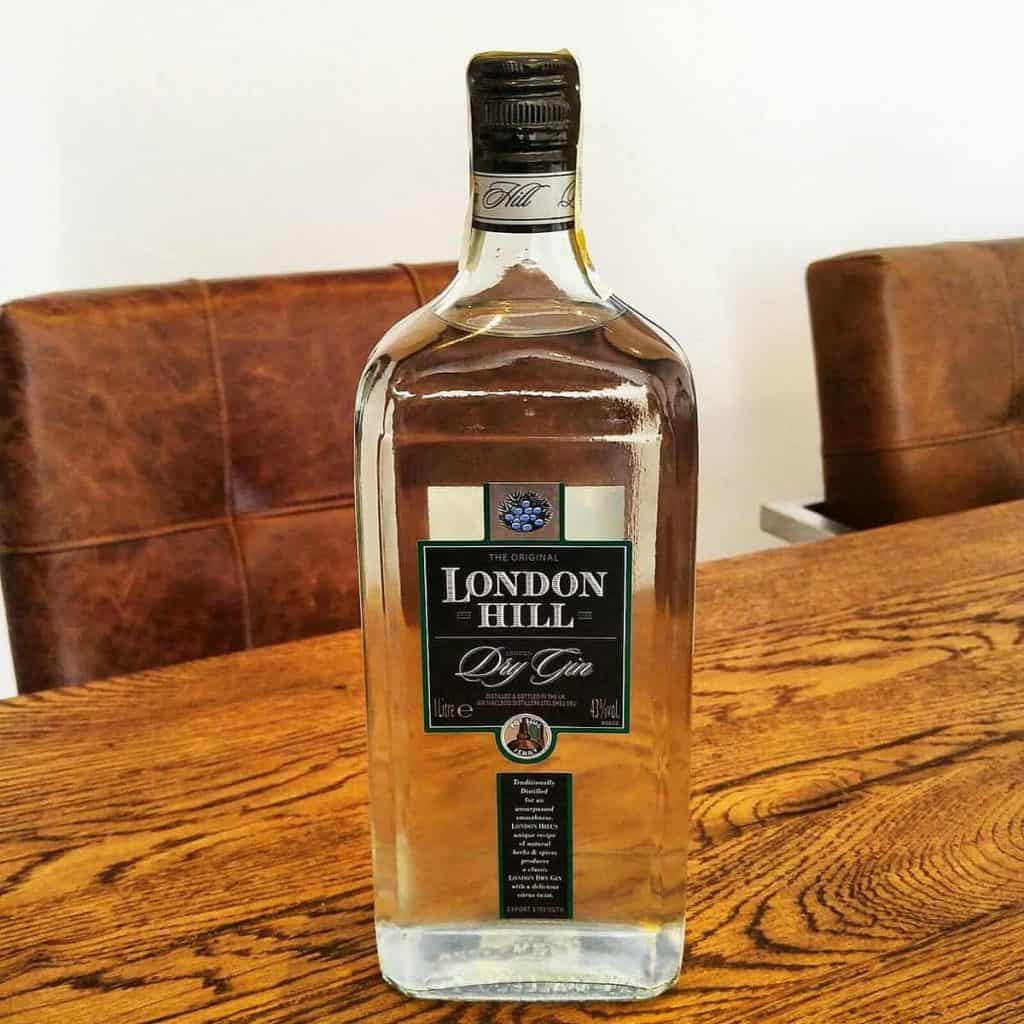 Auf einem Holztisch steht eine große Flasche London Hill Dry Gin. Im Hintergrund sind braune Ledersessel und eine weiße Wand zu sehen.