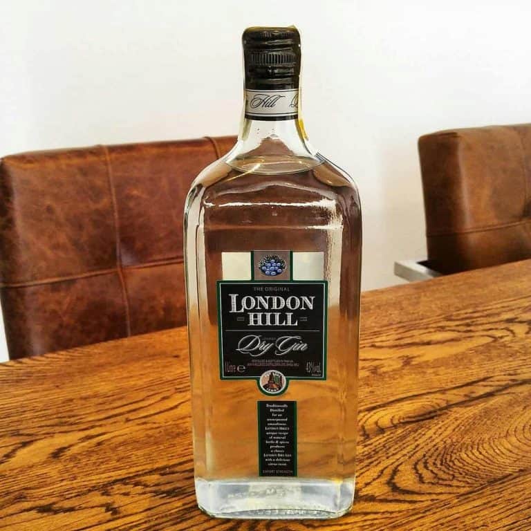 Auf einem Holztisch steht eine große Flasche London Hill Dry Gin. Im Hintergrund sind braune Ledersessel und eine weiße Wand zu sehen.
