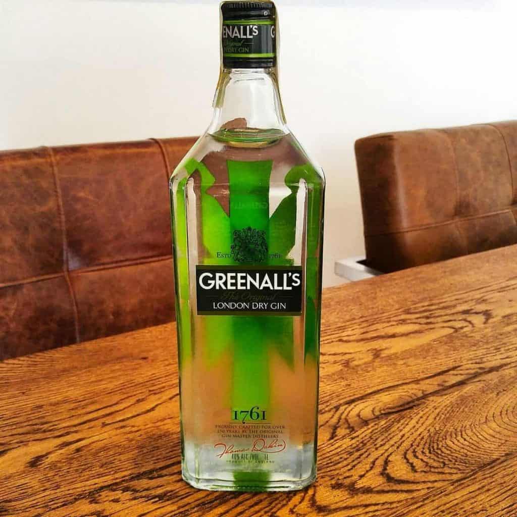 Auf einem Holztisch steht eine Flasche Greenalls London Dry Gin, im Hintergrund sind braune gepolsterte Stühle zu sehen. Die durchsichtige Flasche hat ein grünes Etikett und einen grünen Verschluss, und unten ist die Zahl 1761 aufgedruckt.