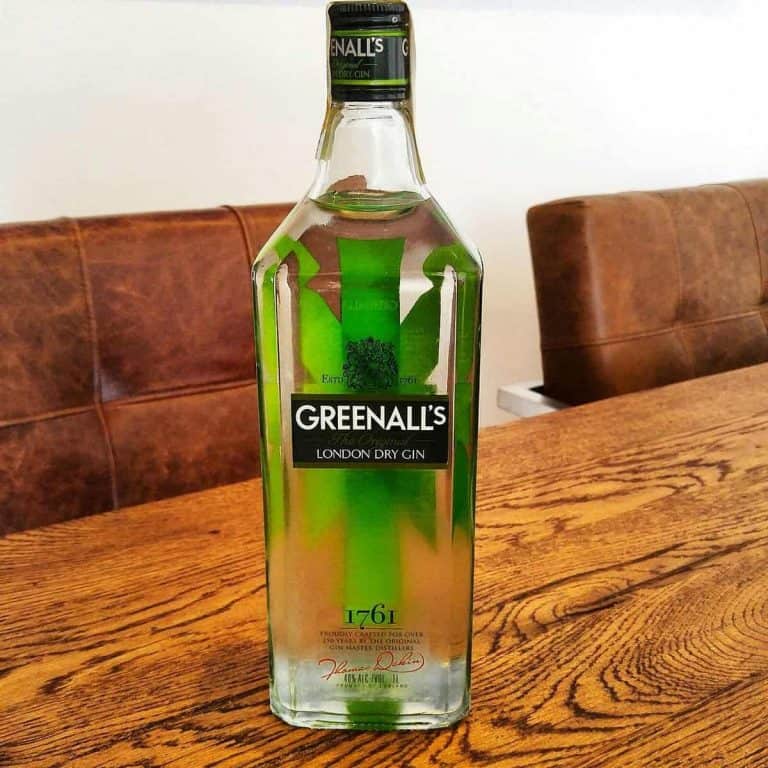 Auf einem Holztisch steht eine Flasche Greenalls London Dry Gin, im Hintergrund sind braune gepolsterte Stühle zu sehen. Die durchsichtige Flasche hat ein grünes Etikett und einen grünen Verschluss, und unten ist die Zahl 1761 aufgedruckt.