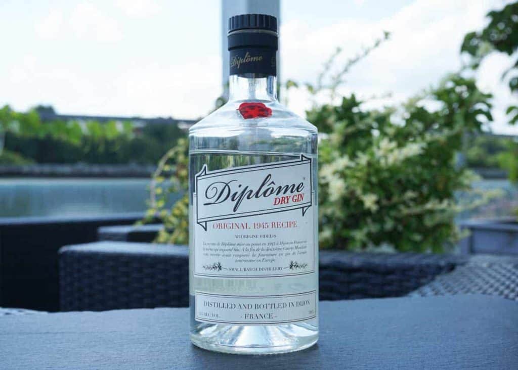 Auf einem dunklen Tisch im Freien steht eine durchsichtige Flasche Diplôme Dry Gin, im Hintergrund sind Pflanzen und ein Gewässer schemenhaft zu erkennen.