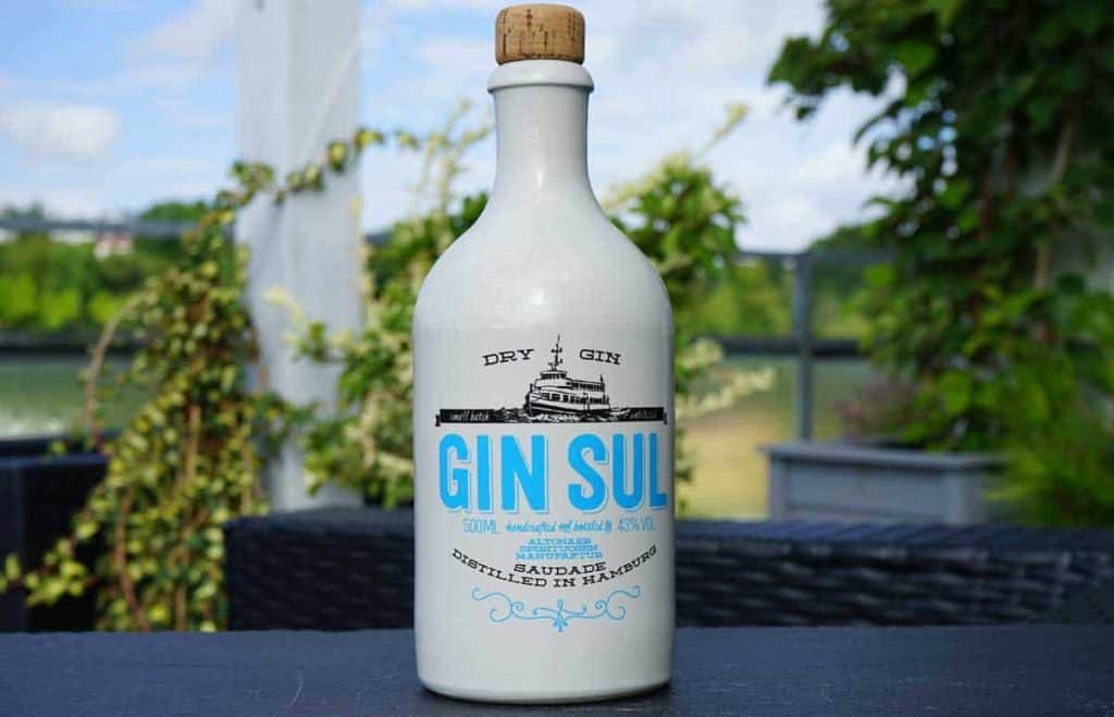 Auf einem Tisch im Freien steht eine weiße Flasche „Gin Sul“ Dry Gin mit Korkverschluss; im unscharfen Hintergrund sind Pflanzen und ein Geländer zu sehen.