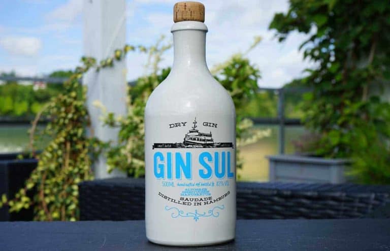 Auf einem Tisch im Freien steht eine weiße Flasche „Gin Sul“ Dry Gin mit Korkverschluss; im unscharfen Hintergrund sind Pflanzen und ein Geländer zu sehen.