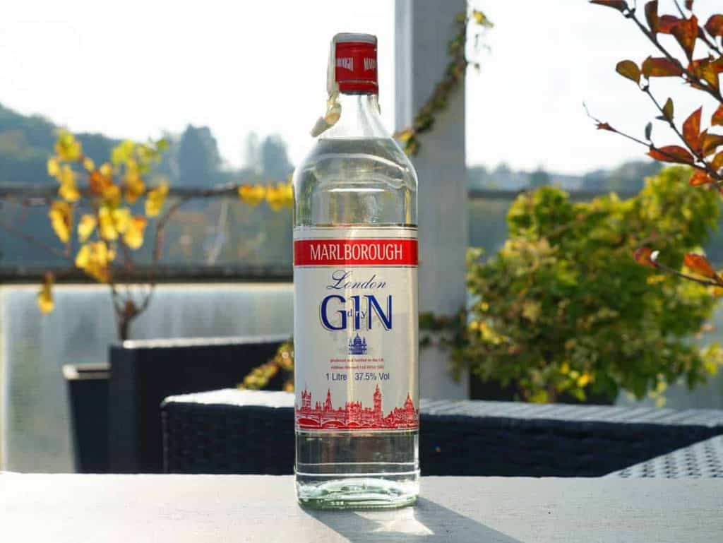 Auf einem Tisch im Freien steht eine Flasche „Marlborough London Gin“, im Hintergrund sind Terrassenmöbel und Herbstlaub zu sehen.