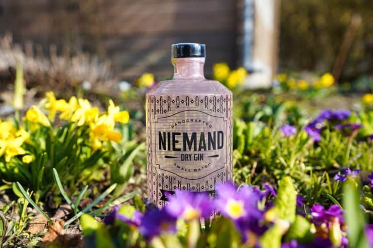 Eine Flasche Niemand Gin steht an einem sonnigen Tag im Freien inmitten blühender gelber und violetter Blumen, der Hintergrund ist unscharf.