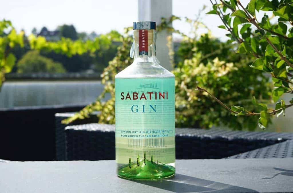 An einem sonnigen Tag steht eine Flasche Sabatini Gin auf einem Tisch im Freien, umgeben von grünen Pflanzen und mit einem See im Hintergrund.