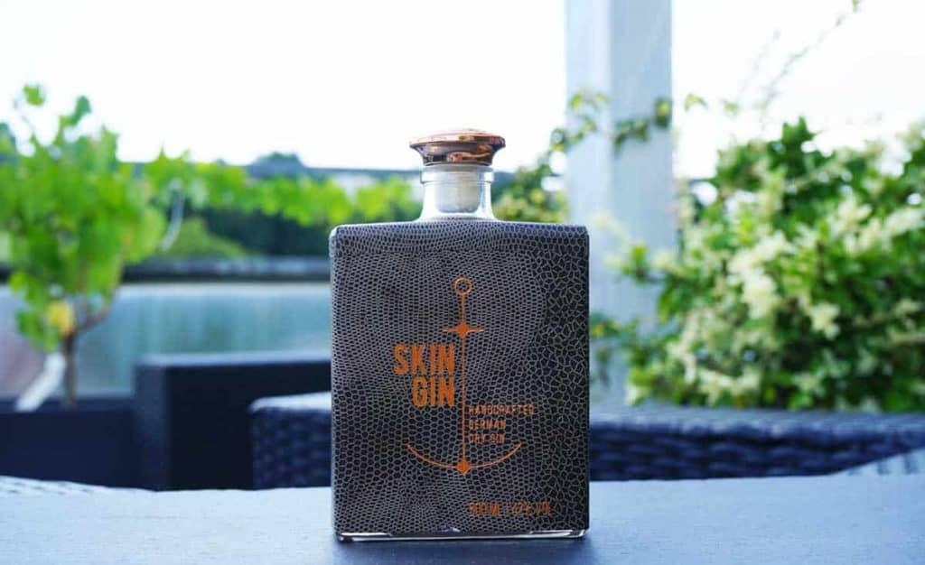 Eine rechteckige Flasche „Skin Gin“ mit einer strukturierten, dunklen Hülle und einem goldenen Anker-Motiv steht auf einem Tisch im Freien, während im Hintergrund Grünpflanzen und Terrassenmöbel verschwommen zu sehen sind.
