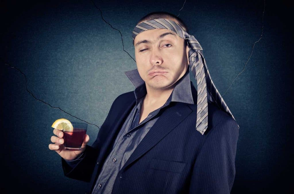 Ein Mann im Anzug, dessen Krawatte locker um den Kopf liegt, hält ein Getränk mit einer Zitronenscheibe in der Hand, während er vor einem dunklen, rissigen Hintergrund zwinkert und eine alberne Grimasse schneidet.