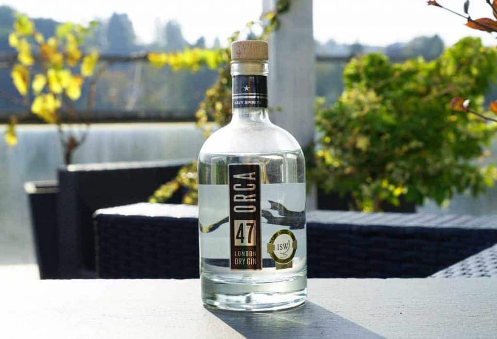 Eine durchsichtige Flasche Orga London Dry Gin steht auf einem Tisch im Freien im Sonnenlicht, im Hintergrund sind Grünpflanzen und schwarze Terrassenmöbel verschwommen zu sehen.