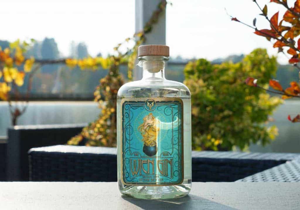 Auf einem Tisch im Freien steht eine durchsichtige Glasflasche mit einem Holzverschluss und einem blauen, verzierten Etikett, umgeben von herbstlichen Pflanzen im sanften Sonnenlicht.
