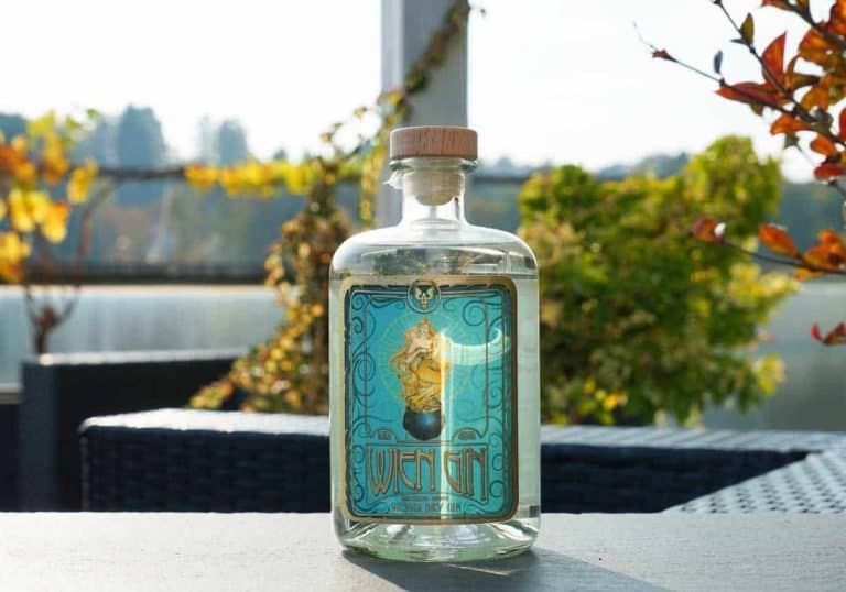 Auf einem Tisch im Freien steht eine durchsichtige Glasflasche mit einem Holzverschluss und einem blauen, verzierten Etikett, umgeben von herbstlichen Pflanzen im sanften Sonnenlicht.